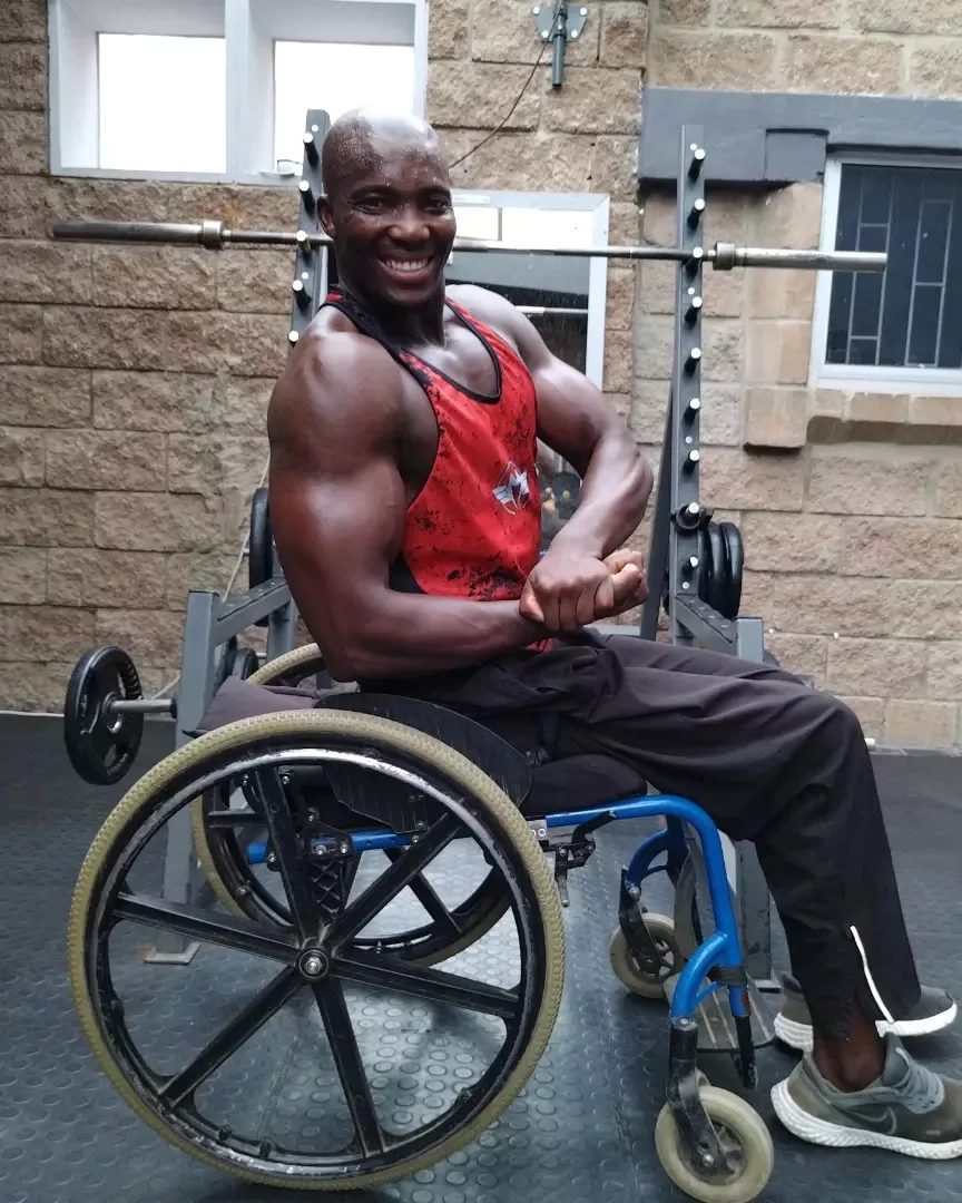 Max Kulati performing front double bicep pose at the gym — red Iron Wolf vest, barbells behind him