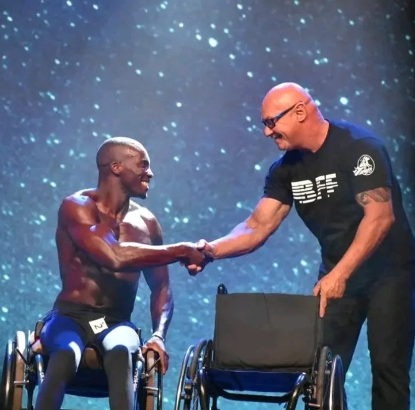 Max Kulati shaking hands with IBFF president on stage — blue sparkle backdrop, Koper Slovenia