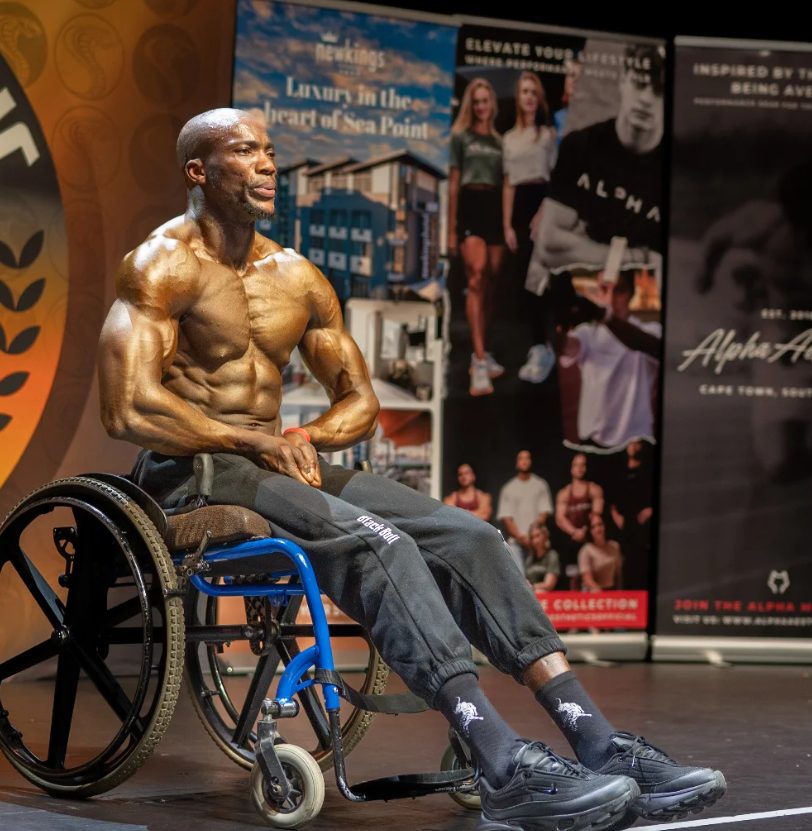 Max Kulati on stage — warm golden lighting, extraordinary muscularity, Black Bull branding